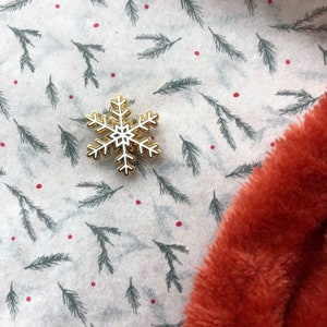 May include: A white snowflake pin with a gold outline on a white background with a green pine needle pattern and red dots. The pin is on a piece of orange faux fur.