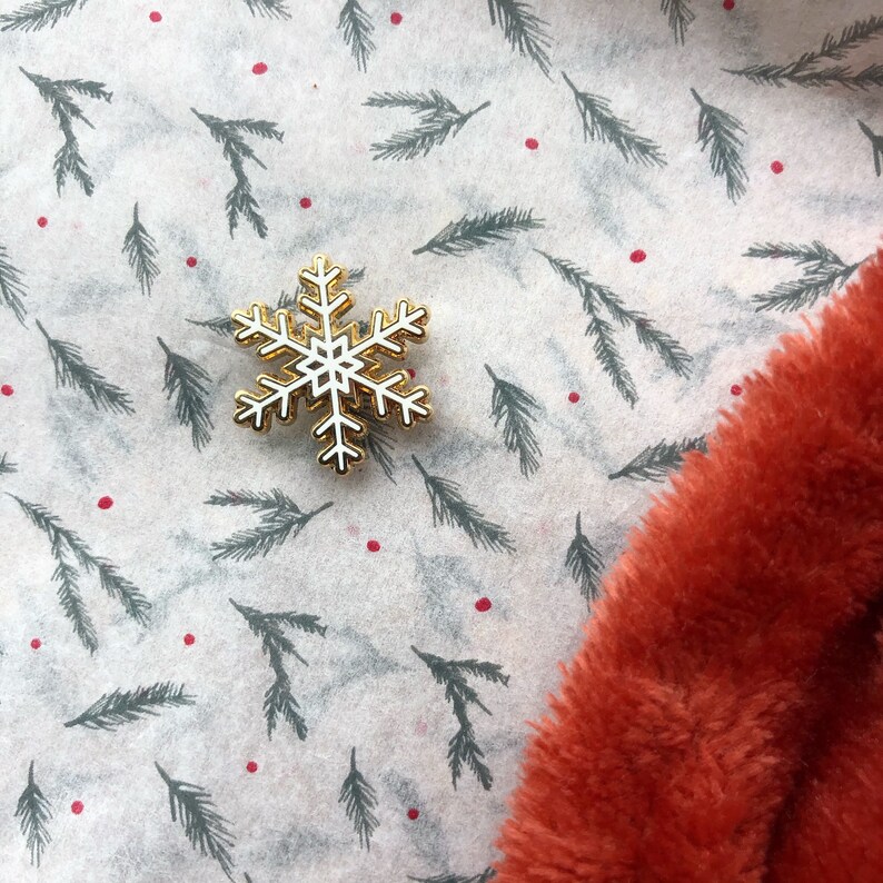 May include: A white snowflake pin with a gold outline on a white background with a green pine needle pattern and red dots. The pin is on a piece of orange faux fur.