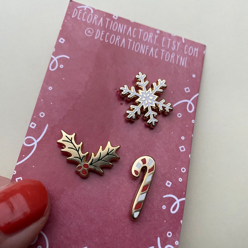 May include: Three gold-colored enamel pins with a Christmas theme. The pins feature a snowflake, a candy cane, and a holly sprig.