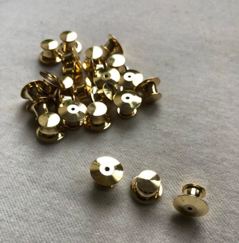 May include: A pile of gold-colored metal pin backs. The pin backs are round with a small hole in the center. There are 18 pin backs in the pile.
