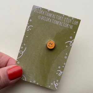Può includere: Uno spillo in smalto con faccina sorridente gialla su una carta verde con coriandoli bianchi e il testo "decorationfactory.etsy.com @decorationfactorynl".