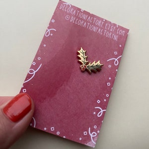 May include: Gold-colored holly leaf pin with red berries. The pin is on a red background with white confetti.