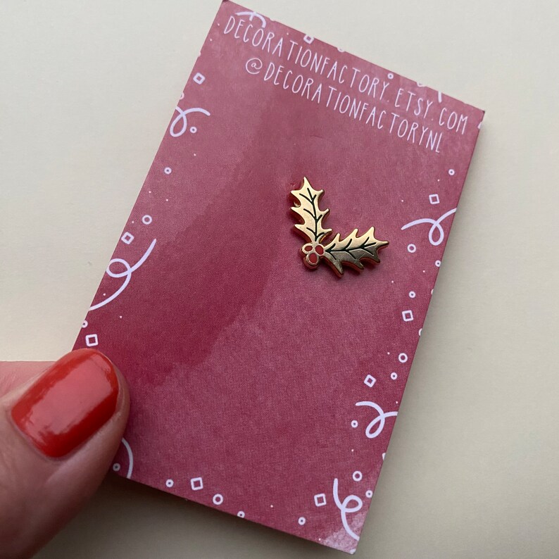 May include: Gold-colored holly leaf pin with red berries. The pin is on a red background with white confetti.