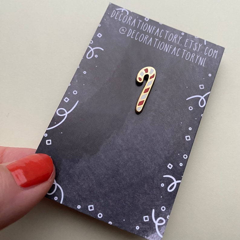 May include: A gold-colored candy cane enamel pin with red and white stripes on a black background with white polka dots and swirls.