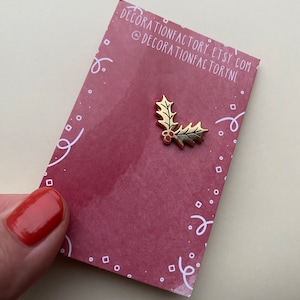 May include: Gold-coloured holly leaf pin with red berries. The pin is on a red background with white confetti.