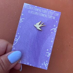 Könnte beinhalten: Eine goldfarbene Anstecknadel mit einer weißen Silhouette eines Vogels im Flug. Die Nadel befindet sich auf einer lila Karte mit weißen Konfetti und dem Text "decorationfactory.etsy.com @decorationfactorynl".