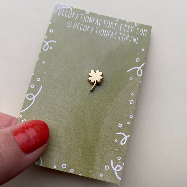 May include: A gold four-leaf clover pin on a green card with white polka dots and a white swirl design. The card has the text "decorationfactory.etsy.com @decorationfactorynl"