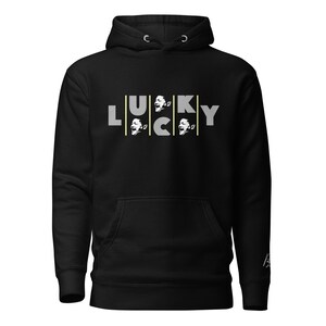 May include: Black hooded sweatshirt with the word "LUCKY" in gray letters. The letters "U", "C", and "K" each have a vertical yellow line and a graphic of a person's face. The sweatshirt has a front pocket and a drawstring hood.