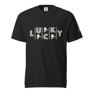 May include: Black t-shirt with the word "LUCKY" in a gray, stylized font. The letters "U" and "C" are formed by vertical yellow lines with a black and white image of a person's face inside each letter. The shirt has short sleeves.