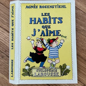 May include: A hardcover book titled "LES HABITS QUE J'AIME" by Agnès Rosenstiehl. The cover features illustrations of two children and the text "CYCLOPÉDIE LAROUSSE". The book has a yellow background with blue and black text.