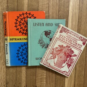 May include: Three vintage books: one orange and blue with fleur-de-lis patterns titled Speaking French, one light green with a globe illustration titled Listen and Sing, and one cream-colored with red floral and bird illustrations titled Histoire d'un Cerf-Volant et d'un Parapluie.