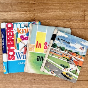 May include: A stack of vintage children's books with colorful covers. Titles include "Sombrero," "In School," and "At School." The books are arranged on a wooden surface, showcasing their retro design and illustrations.