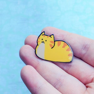May include: A yellow and orange enamel pin of a cartoon cat curled up in a sleeping position.