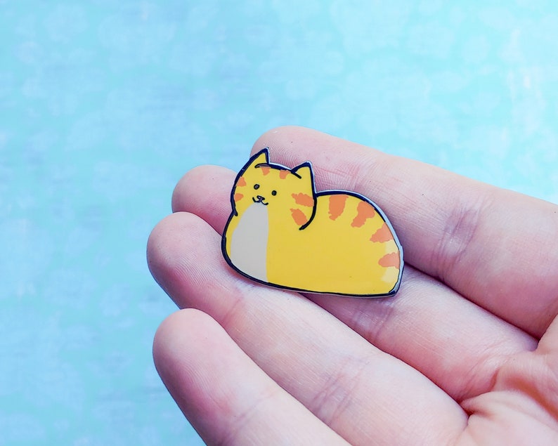 May include: A yellow and orange enamel pin of a cartoon cat curled up in a sleeping position.