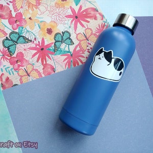 Puede incluir: Una botella de agua de acero inoxidable azul con una pegatina de gato blanco. La pegatina muestra un gato blanco y negro con una expresi&oacute;n linda.