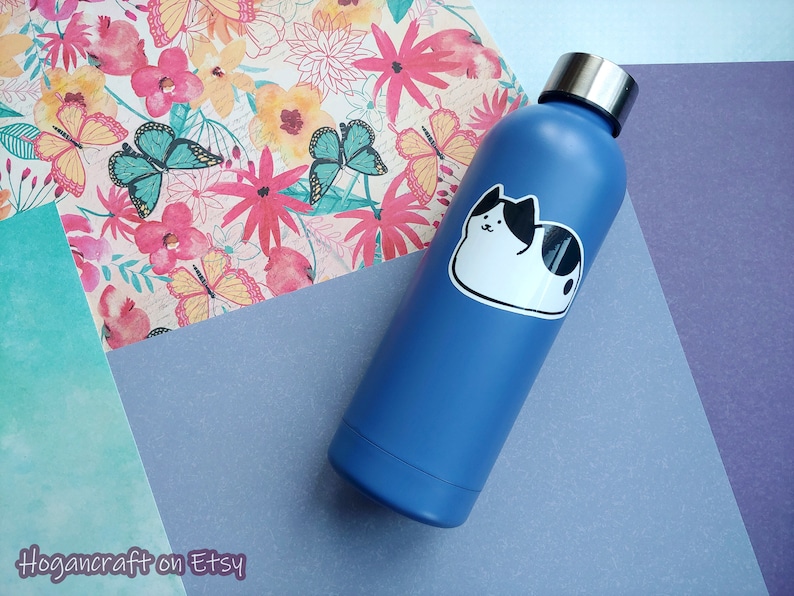 Puede incluir: Una botella de agua de acero inoxidable azul con una pegatina de gato blanco. La pegatina muestra un gato blanco y negro con una expresi&oacute;n linda.