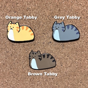 May include: Three enamel pins featuring cartoon cats in different colors. The pins are labeled "Orange Tabby", "Grey Tabby", and "Brown Tabby".