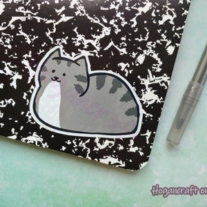 Puede incluir: Una pegatina de gato de dibujos animados gris y blanco sobre un fondo moteado en blanco y negro. La pegatina es de un gato acostado de lado con las patas metidas debajo del cuerpo.