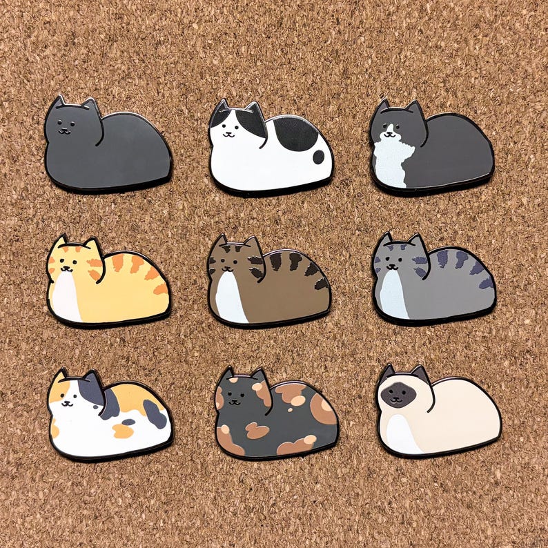 May include: Nine enamel pins featuring different colored cats in a curled up sleeping position. The cats are all different colors and patterns, including black, white, orange, brown, gray, and calico.