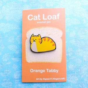 May include: A white enamel pin shaped like a slice of bread with an orange tabby cat curled up inside. The pin says "Cat Loaf" and "Orange Tabby" and is by Alyssa H (Hogancraft).