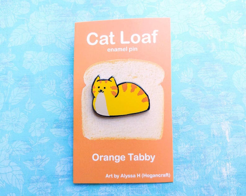 May include: A white enamel pin shaped like a slice of bread with an orange tabby cat curled up inside. The pin says "Cat Loaf" and "Orange Tabby" and is by Alyssa H (Hogancraft).
