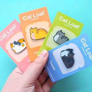 May include: Four enamel pins featuring cats curled up in a loaf shape on a slice of bread. The pins are on cards with the text "Cat Loaf enamel pin" and the cat's breed name. The breeds are Orange Tabby, Tuxedo, Calico, and Gray Tabby.
