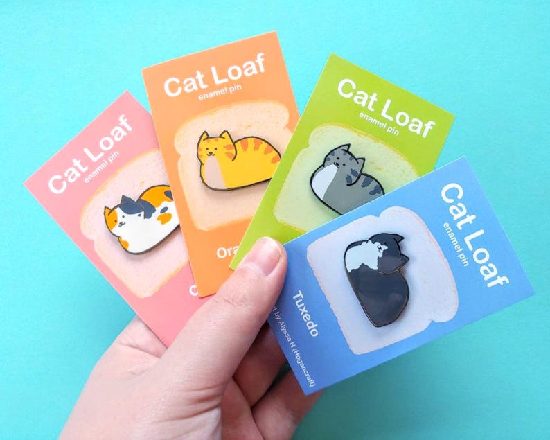 May include: Four enamel pins featuring cats curled up in a loaf shape on a slice of bread. The pins are on cards with the text "Cat Loaf enamel pin" and the cat's breed name. The breeds are Orange Tabby, Tuxedo, Calico, and Gray Tabby.