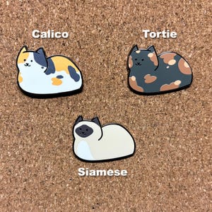 May include: Three enamel pins featuring different cat breeds. The pins are in the shape of cats and are labeled "Calico", "Tortie", and "Siamese".
