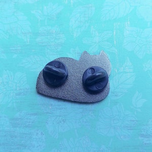 May include: The back of a silver enamel pin featuring a cartoon cat laying down. The pin has two black metal clasps on the back.