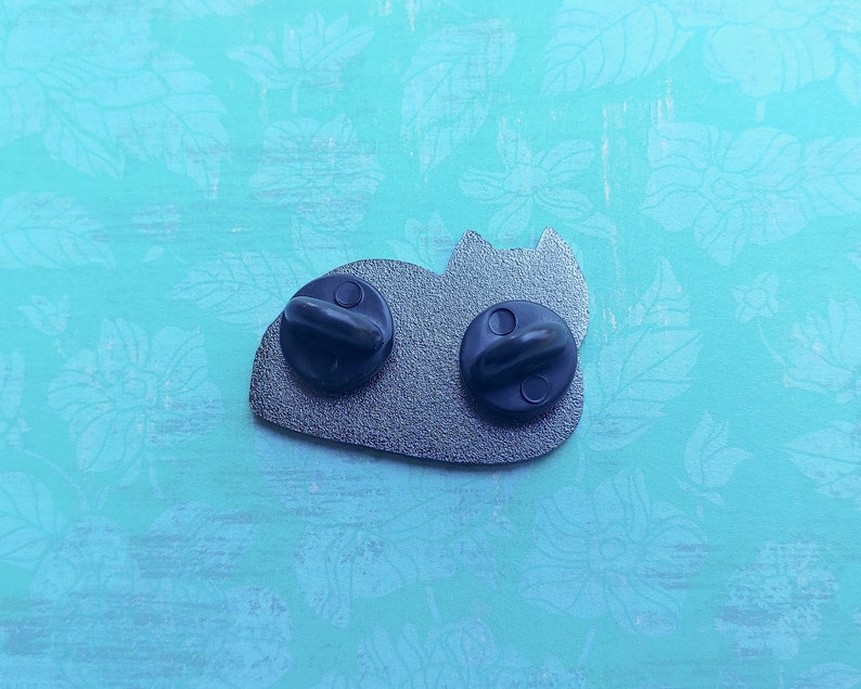 May include: The back of a silver enamel pin featuring a cartoon cat laying down. The pin has two black metal clasps on the back.