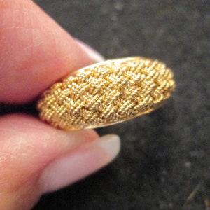 May include: A gold ring with a textured, woven design. The ring is held between fingers, highlighting its intricate pattern and metallic sheen. The ring's surface features a detailed, repeating pattern, giving it a unique and eye-catching appearance.