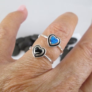 May include: Two silver rings with heart-shaped settings. One ring has a black stone in the heart, and the other has a blue opal.