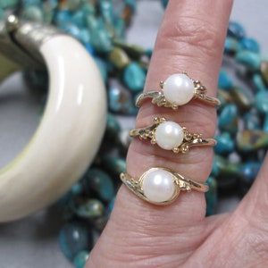 May include: Three gold rings with white pearl stones. The rings are set on a finger with a turquoise background.