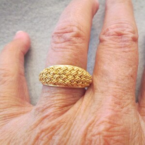 May include: A gold-coloured ring with a woven design is displayed on a person's finger. The ring has a rounded shape and a textured surface, showcasing a detailed pattern. The ring is a piece of jewellery.