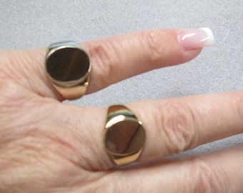 May include: Two gold signet rings with oval-shaped tops. The rings are worn on the fingers of a hand.
