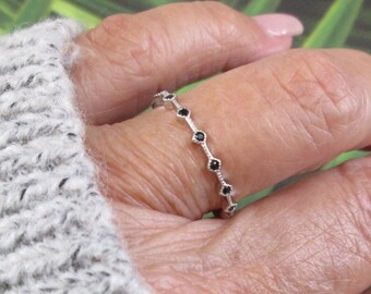 May include: A silver ring with small black gemstones set in a delicate design. The ring is worn on a finger, with a gray sweater sleeve visible in the background. The ring's design features a series of small, dark stones.