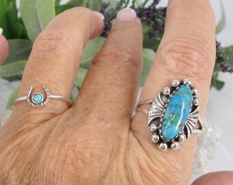 May include: Two silver rings with turquoise stones. The larger ring has an oval shaped turquoise stone with a silver setting. The smaller ring has a round turquoise stone set in a horseshoe shape.