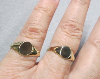 May include: Two gold signet rings with oval tops on a person's hand. The rings are worn on the index and middle fingers.