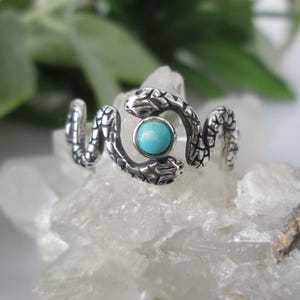 May include: A silver-toned ring featuring a snake design with a turquoise stone center. The ring's snakes are detailed with textured scales. The turquoise stone is a bright, round shape. The ring is displayed on a white, crystalline surface.