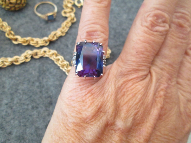 May include: A silver ring featuring a large, rectangular, purple gemstone. The gemstone has a gradient of colour from dark purple to light purple. The ring is set with smaller, clear gemstones around the main stone.