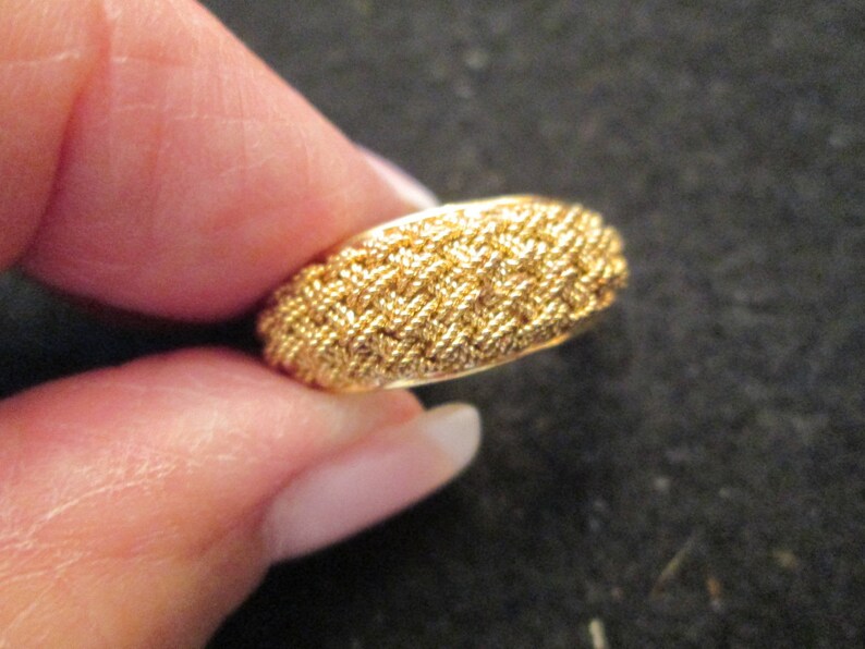May include: A gold ring with a textured, woven design. The ring is held between fingers, highlighting its intricate pattern and metallic sheen. The ring's surface features a detailed, repeating pattern, giving it a unique and eye-catching appearance.