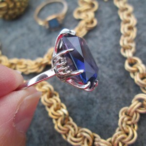 May include: A silver ring featuring a large, faceted, purple gemstone set in a claw setting. The ring is surrounded by a gold chain.