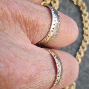 May include: Two gold wedding bands with a textured design. The bands are worn on a person's fingers.