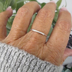 May include: A silver ring with a hammered bar design. The ring is on a finger, with a gray sweater in the background. The ring's band has a twisted rope design.