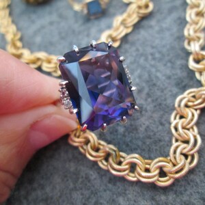 May include: A gold chain necklace with a large, faceted, purple gemstone ring. The ring has small, clear gemstones around the setting.