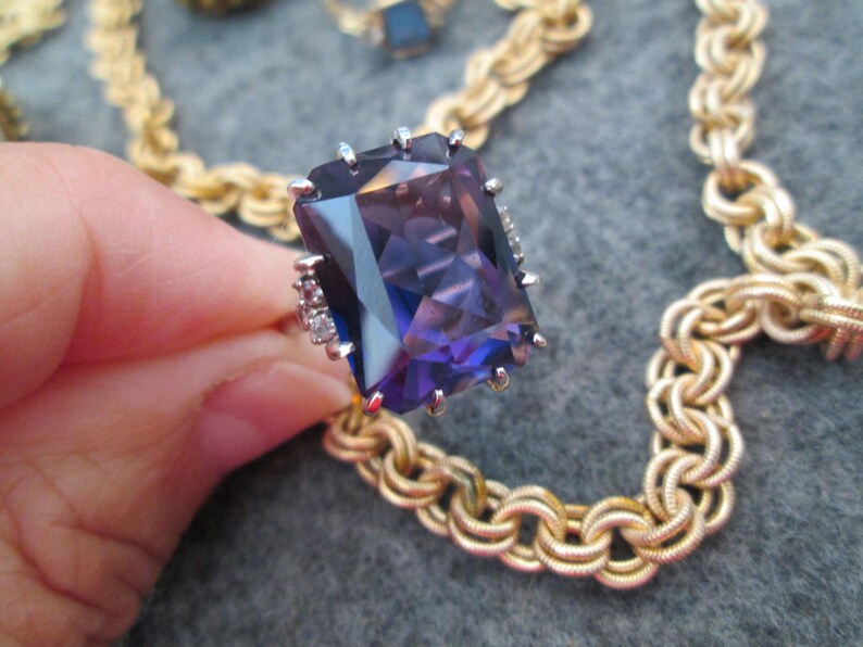 May include: A gold chain necklace with a large, faceted, purple gemstone ring. The ring has small, clear gemstones around the setting.