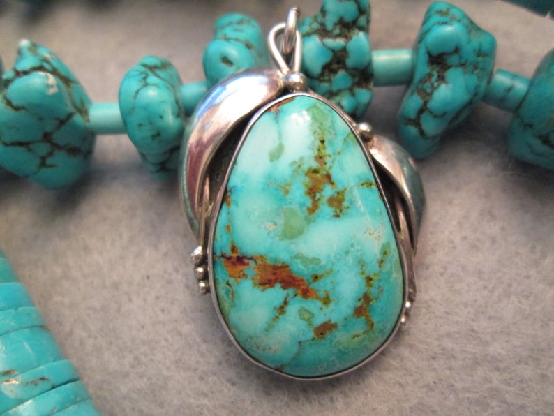 Peut inclure: Un pendentif en turquoise avec un sertissage en argent. La turquoise est de couleur bleu clair avec des veines brunes et un motif marbr&eacute;. Le pendentif est entour&eacute; de perles en turquoise.