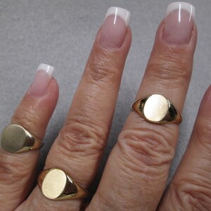 May include: Three gold signet rings with oval-shaped tops. The rings are worn on different fingers of a hand.