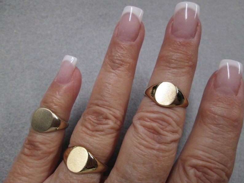 May include: Three gold signet rings with oval-shaped tops. The rings are worn on different fingers of a hand.