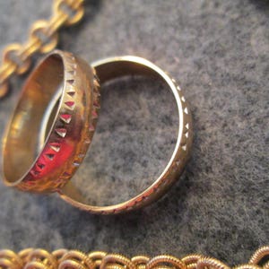 May include: Two gold-colored rings with a textured design. The ring on the left has a more intricate design with small triangles. The ring on the right has a smooth, rounded design.